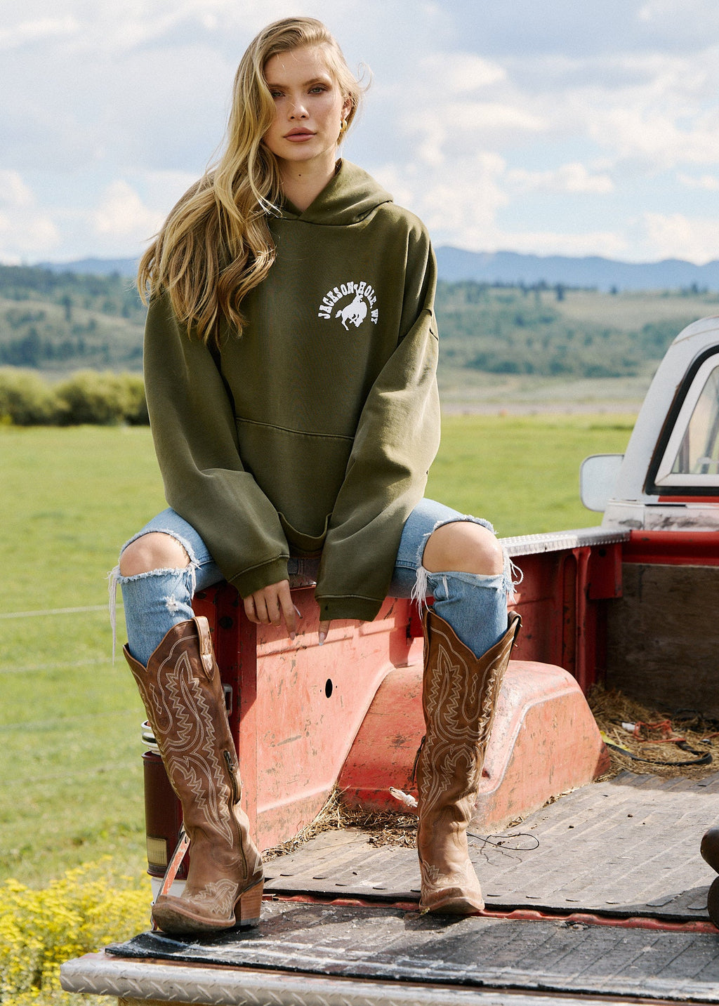 Army Green | Woman in a green hoodie and cowboy boots sitting on a truck bed with a scenic background